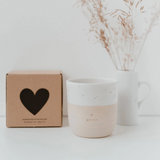 Stillleben auf einem weißen Tisch: Ein Eulenschnitt Keramikbecher "Mama" (weiß/beige), eine dazu passende Eulenschnitt Geschenkbox aus Kraftpapier mit einem schwarzen Herz, und ein Trockenblumenstrauß in einer weißen Vase.