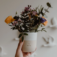Eine Hand hält einen weiß-beigen Keramikbecher der Marke Eulenschnitt, der als Vase für einen bunten Wiesenblumenstrauß mit Rosen und Eukalyptus dient. Gut sichtbare Gravur "Mama" auf dem Becher.