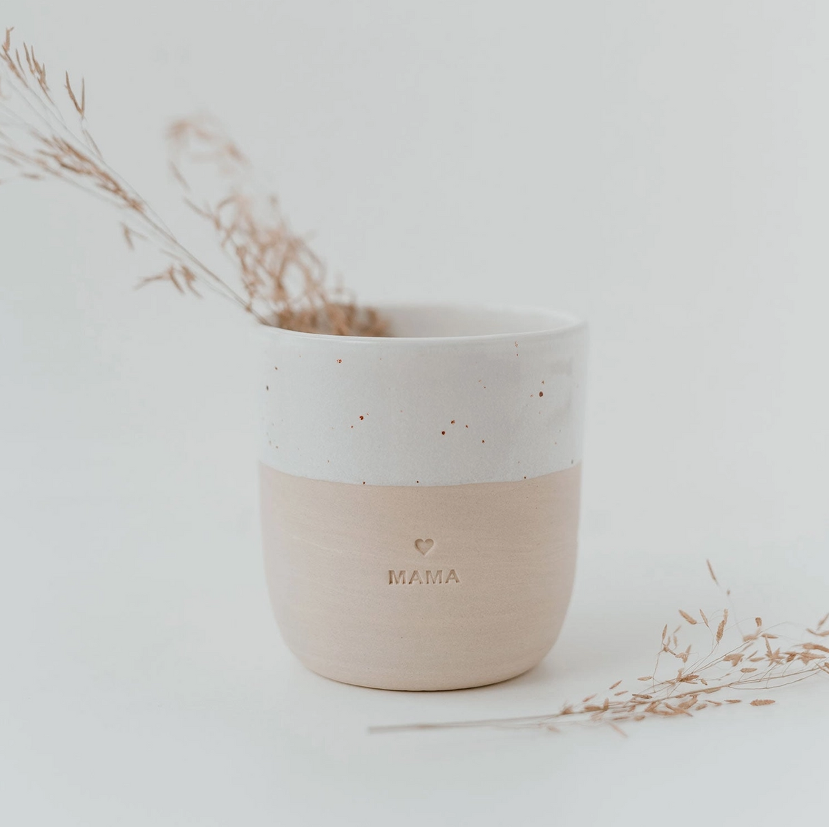 Alleinstehender Eulenschnitt Keramikbecher "Mama" (weiß/beige) auf einem weißen Untergrund, gefüllt mit zarten, getrockneten Gräsern. Minimalistische Boho-Wohnkultur.